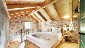 Une chambre avec un mur en pierre, des poutres apparentes en bois au plafond, un lit recouvert de draps blancs et un plancher en bois.