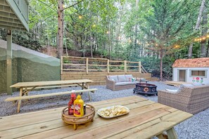 Une table de pique-nique en bois avec un panier de chips et des condiments, un canapé en osier et une petite cheminée d’extérieur.