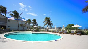 A swimming pool with lounge chairs and umbrellas, surrounded by palm trees and a building with a blue roof. - Apartment sea view Orient bay beach (Saint-Martin)