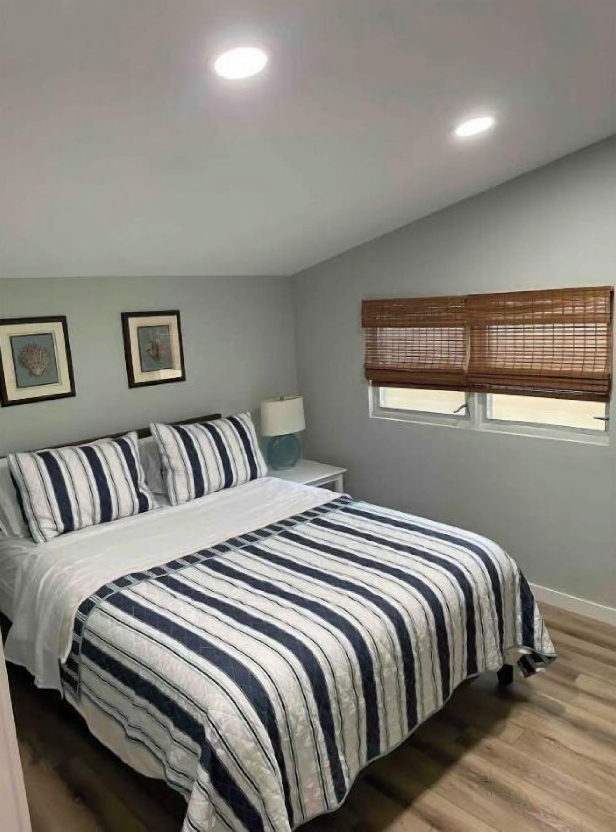 Un dormitorio con cama, mesita de noche, ventana con persianas y dos cuadros en la pared.