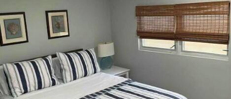 Un dormitorio con cama, mesita de noche, ventana con persianas y dos cuadros en la pared.
