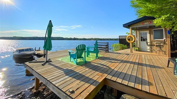A wooden deck with chairs and a table, a small building with a window, and a boat docked by the water.
