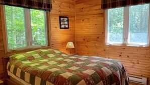 A wooden cabin bedroom with a bed, a ceiling fan, and two windows with curtains.