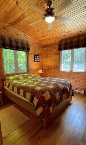 A wooden cabin bedroom with a bed, two windows, a ceiling fan, and a small table with a lamp.