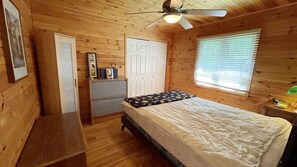 Una habitación con una cama, ventilador de techo, ventana con persianas, una puerta y una pequeña mesa.