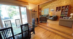 A cozy living room with a wooden ceiling, a glass dining table with chairs, a sofa, a bookshelf, and a large window offering a view of the outdoors.