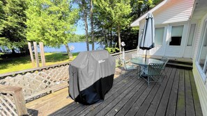 A covered barbecue grill on a wooden deck with a table and chairs, overlooking a lake and a white house.