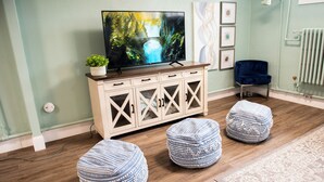 A living room with a television, wooden cabinet, ottomans, and a radiator. - McKinley Mansion Half Building, sleeps 35, Gym, Arcade Room (Pocatello)