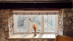 Ein Zimmer mit einer Steinsäule, einem Fenster mit Rollläden, einer Vase mit Trockenpflanzen und einem braunen Stuhl.