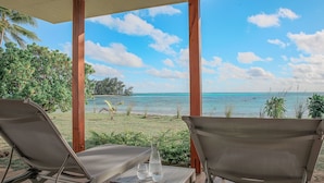 A covered outdoor seating area with two lounge chairs and a table, overlooking a beach with clear blue water and a grassy area with palm trees. - Niu Beach Hotel Moorea (Moorea-Maiao)