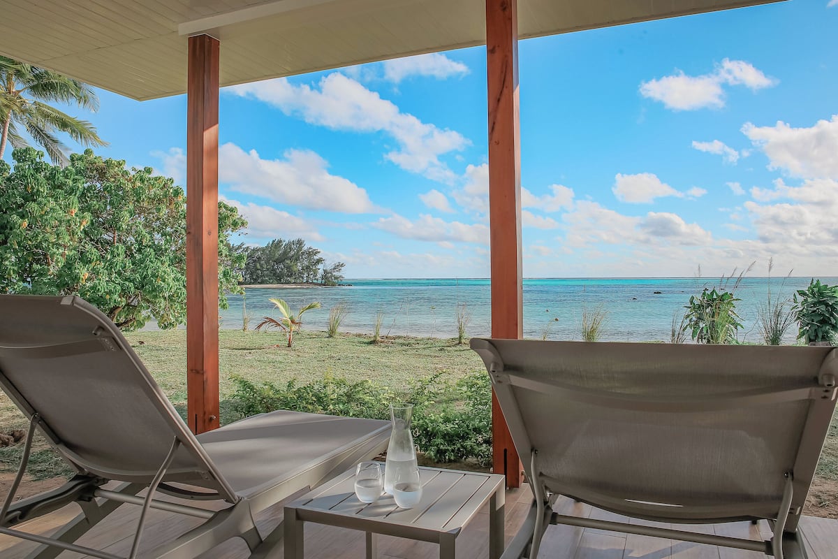 A covered outdoor seating area with two lounge chairs and a table, overlooking a beach with clear blue water and a grassy area with palm trees.