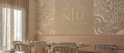 Restaurante com cadeiras de vime, mesas postas com pratos, copos e talheres. A parede apresenta um logotipo grande e padrões decorativos.