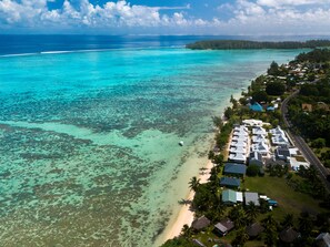 An aerial view of a coastal resort with multiple bungalow-style buildings, a beach, and clear turquoise waters.