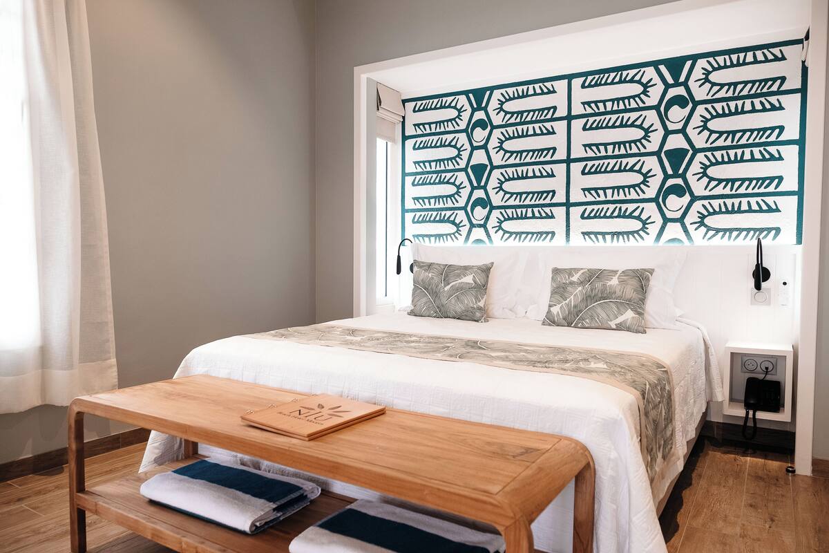 A neatly arranged hotel room with a bed, a wooden bedside table with a book, a nightstand, and a wall-mounted phone.