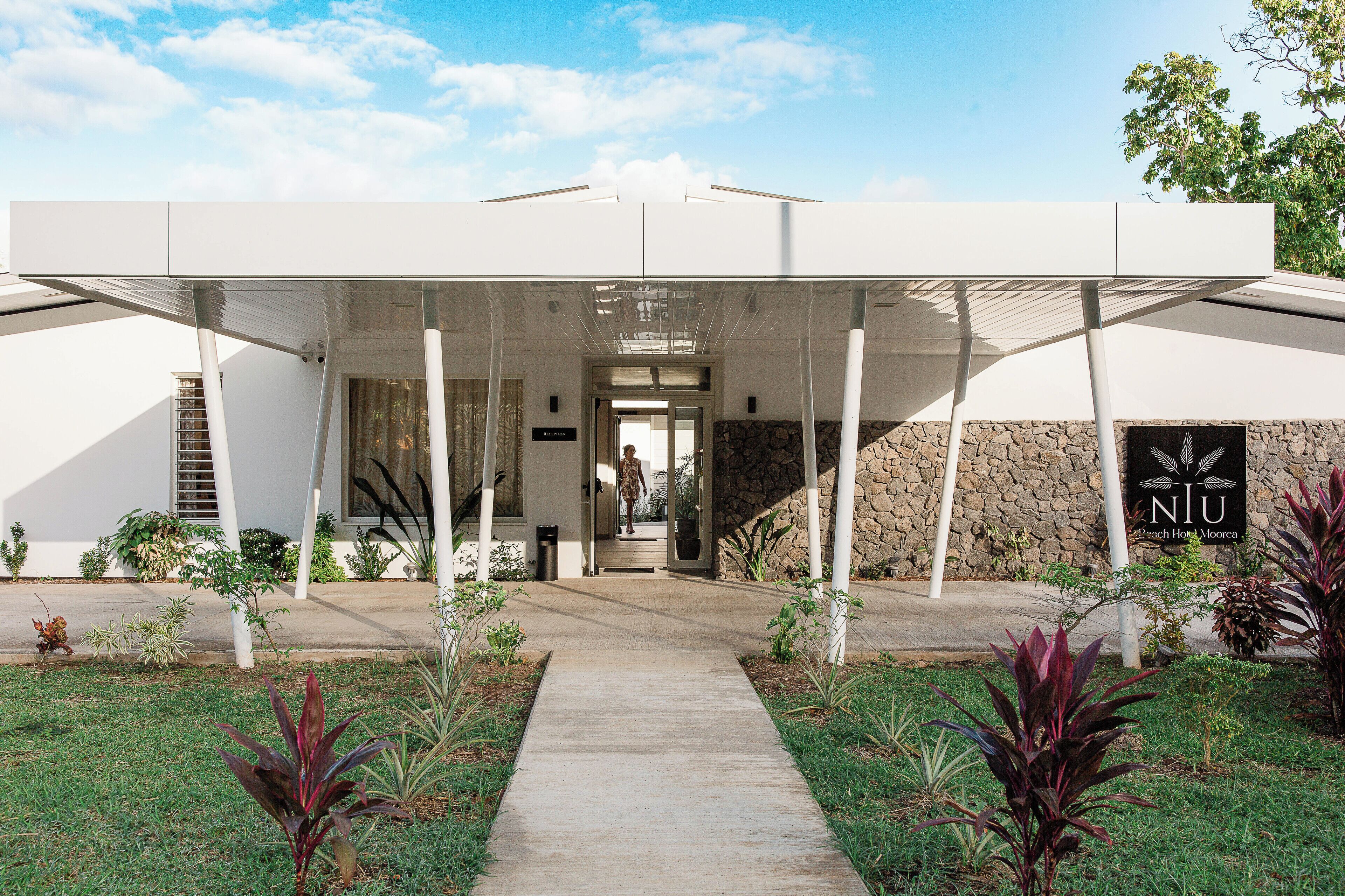 Un edificio moderno con entrada cubierta, pared de piedra y un letrero que dice "NIU Beach House Moraea".