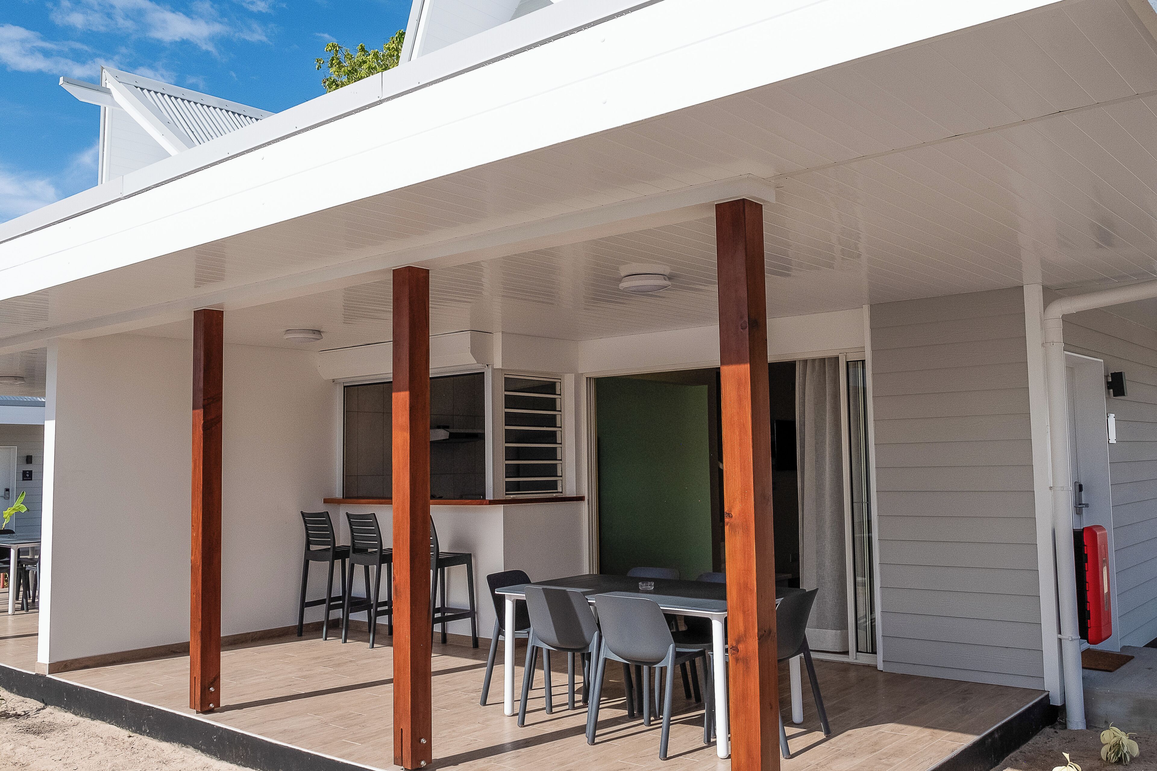 An outdoor covered patio with a dining table and chairs, wooden posts, and a green wall.