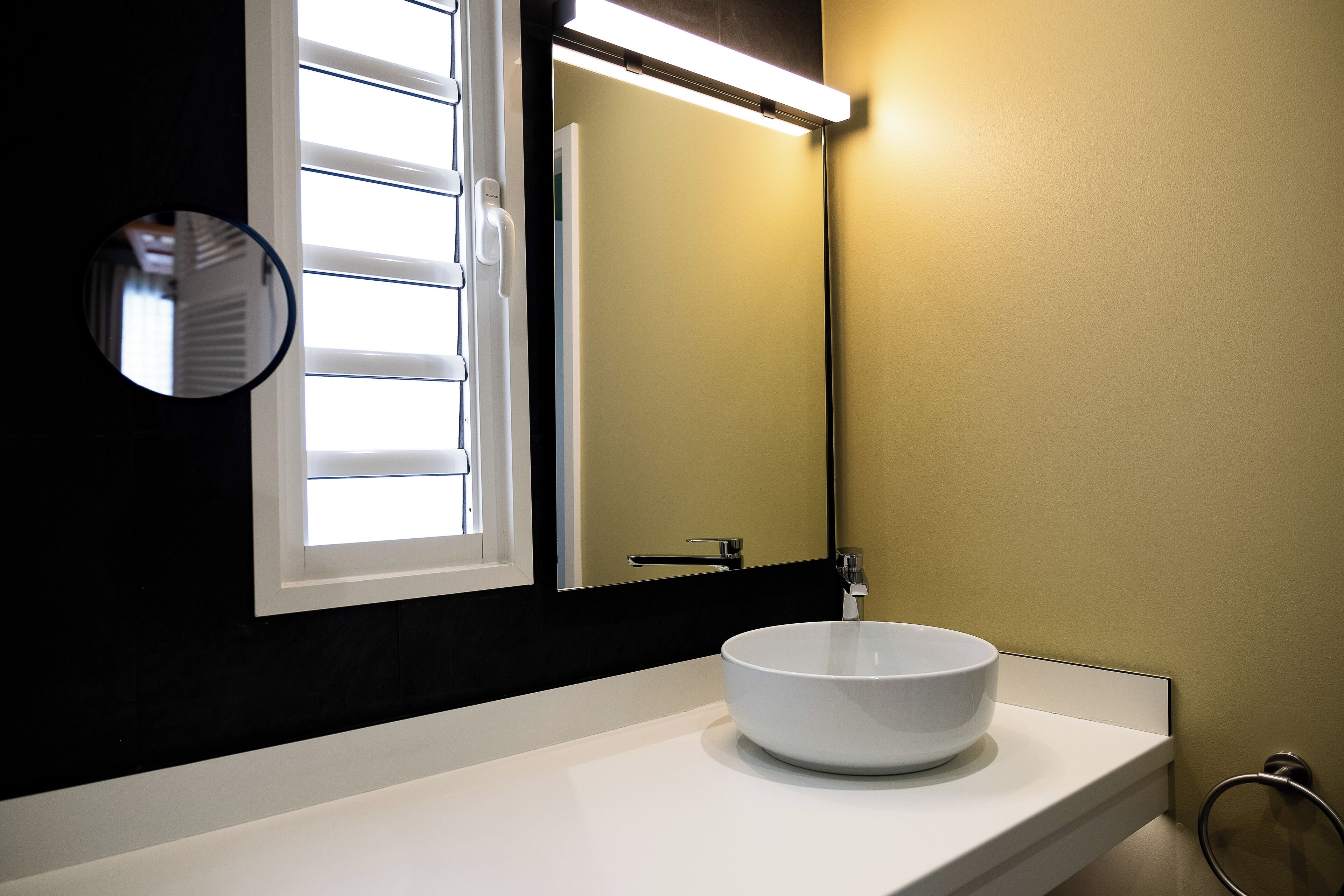 Un baño con lavamanos blanco, espejo y ventana con persianas.