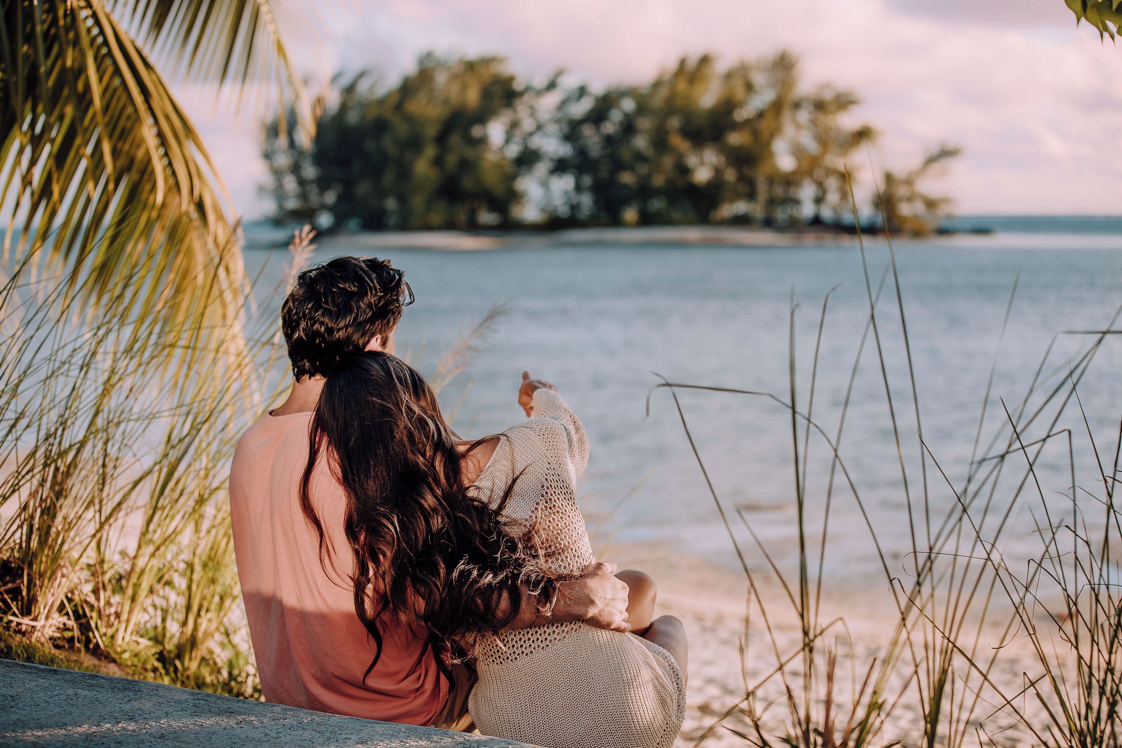 Zwei Personen sitzen auf einer Bank am Strand und blicken auf das Meer und eine entfernte Insel.