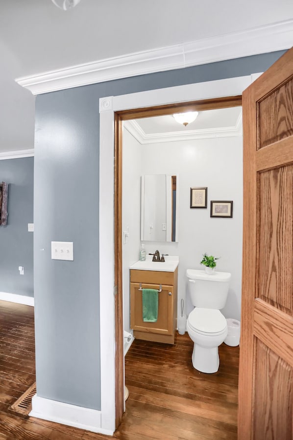A bathroom with a wooden floor, white walls, a white toilet, a sink with a mirror, and a green towel hanging on the door.