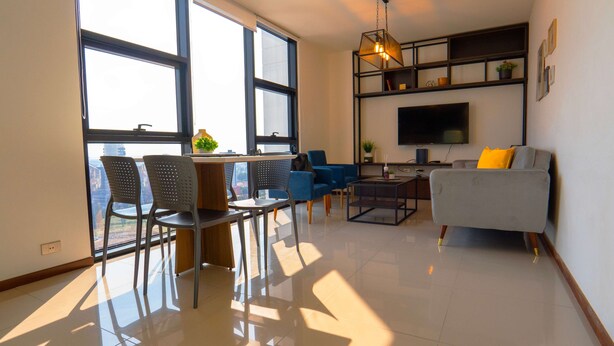 A modern living room with a sofa, chairs, a coffee table, and a TV. There are large windows providing natural light.