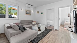 Una sala moderna con un sofá seccional gris, una mesa de centro de vidrio y una alfombra negra. Hay una puerta blanca que da a otra habitación y una ventana con vistas a un edificio y árboles.