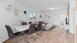 Un comedor moderno con una mesa blanca y sillas oscuras, un sofá y un televisor al fondo.