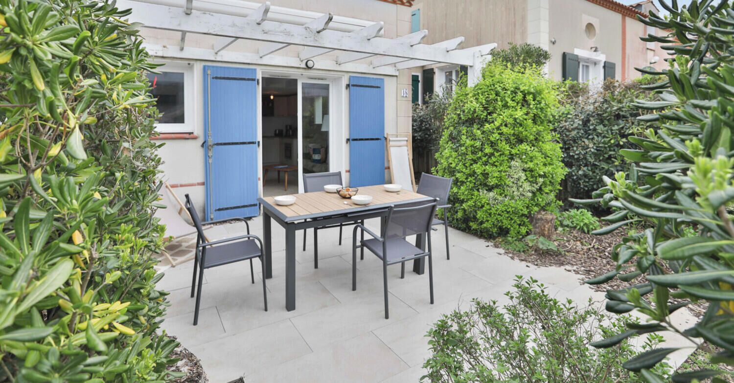 A patio with a table and chairs, surrounded by greenery and a building with blue shutters.