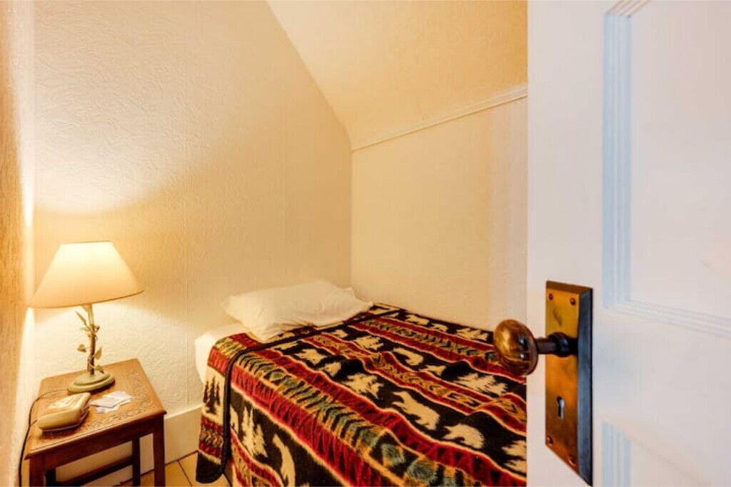 Um quarto pequeno e acolhedor com uma cama de solteiro, uma mesa de cabeceira com abajur e uma porta com puxador redondo.