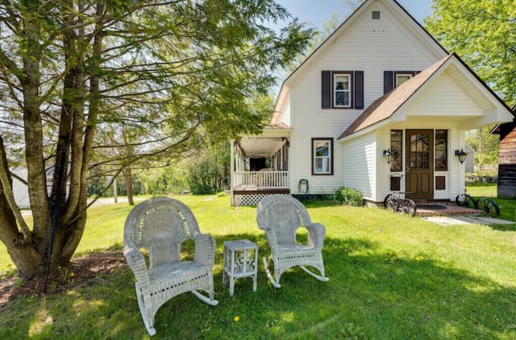 A white house with a porch, two wicker chairs, and a tree.