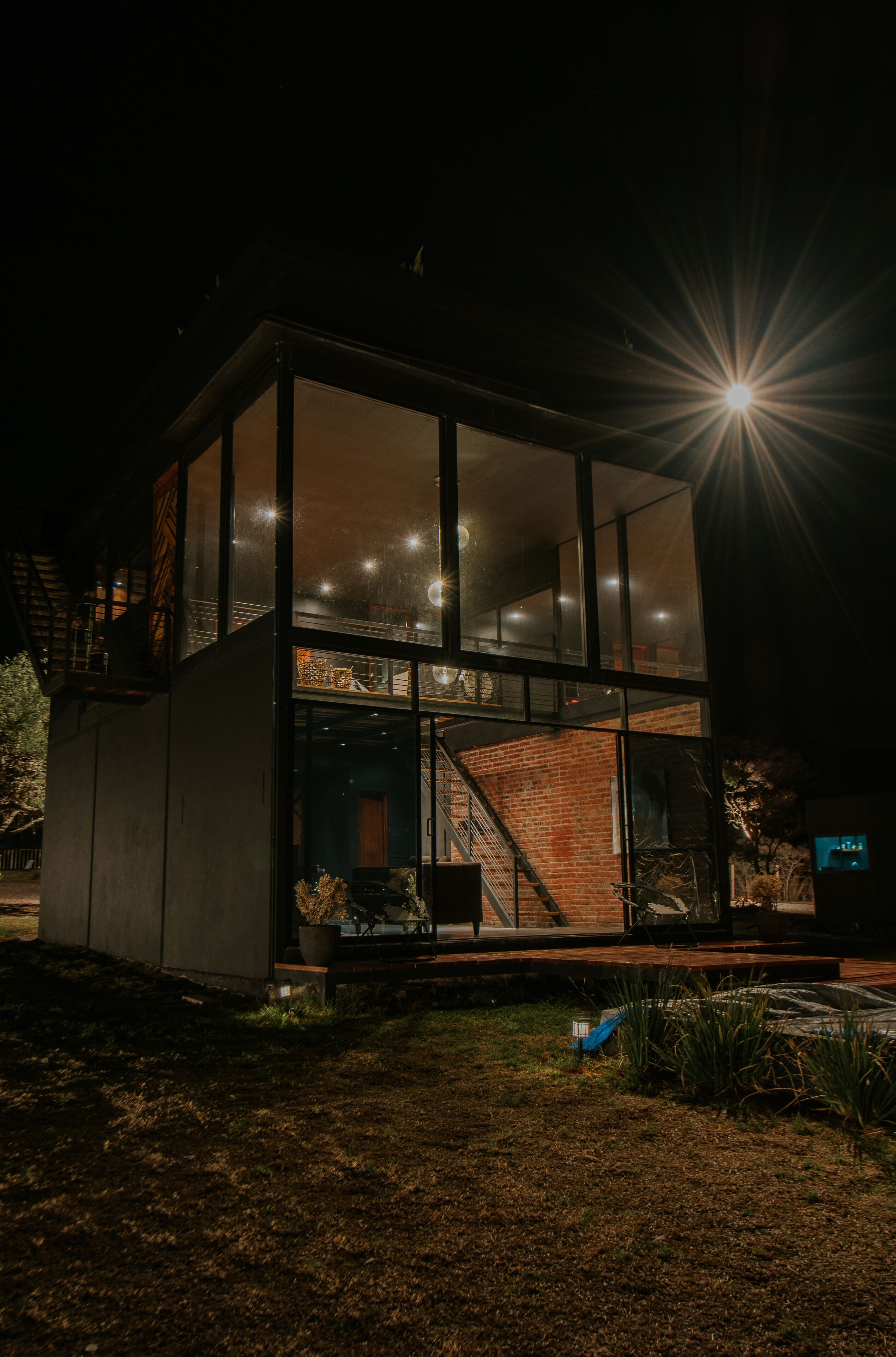A modern glass house with a brick interior, a staircase, and a small outdoor seating area.