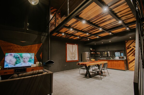 A modern kitchen with a wooden ceiling, a dining area with a table and chairs, and a television in the foreground. - K NAJ Community (San Miguel de Allende)