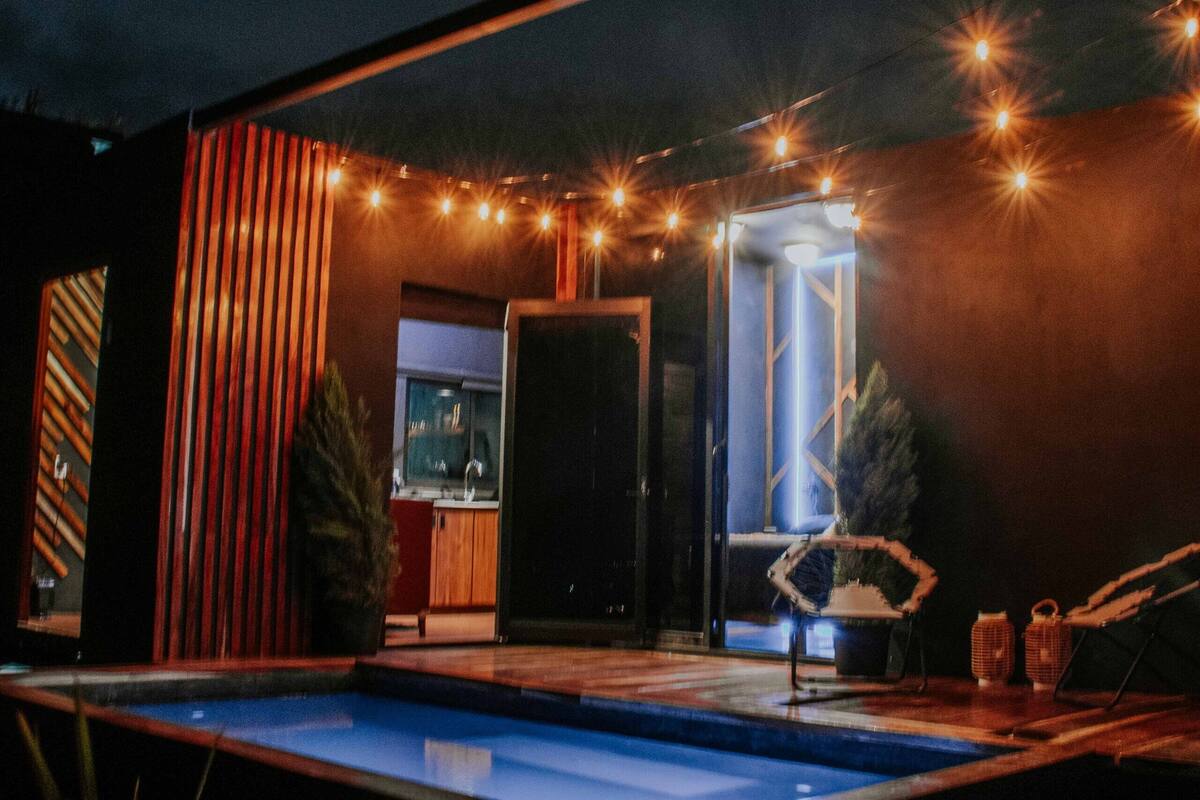 Un espace piscine extérieur moderne, doté d’une structure en verre, de guirlandes lumineuses et d’une terrasse en bois.