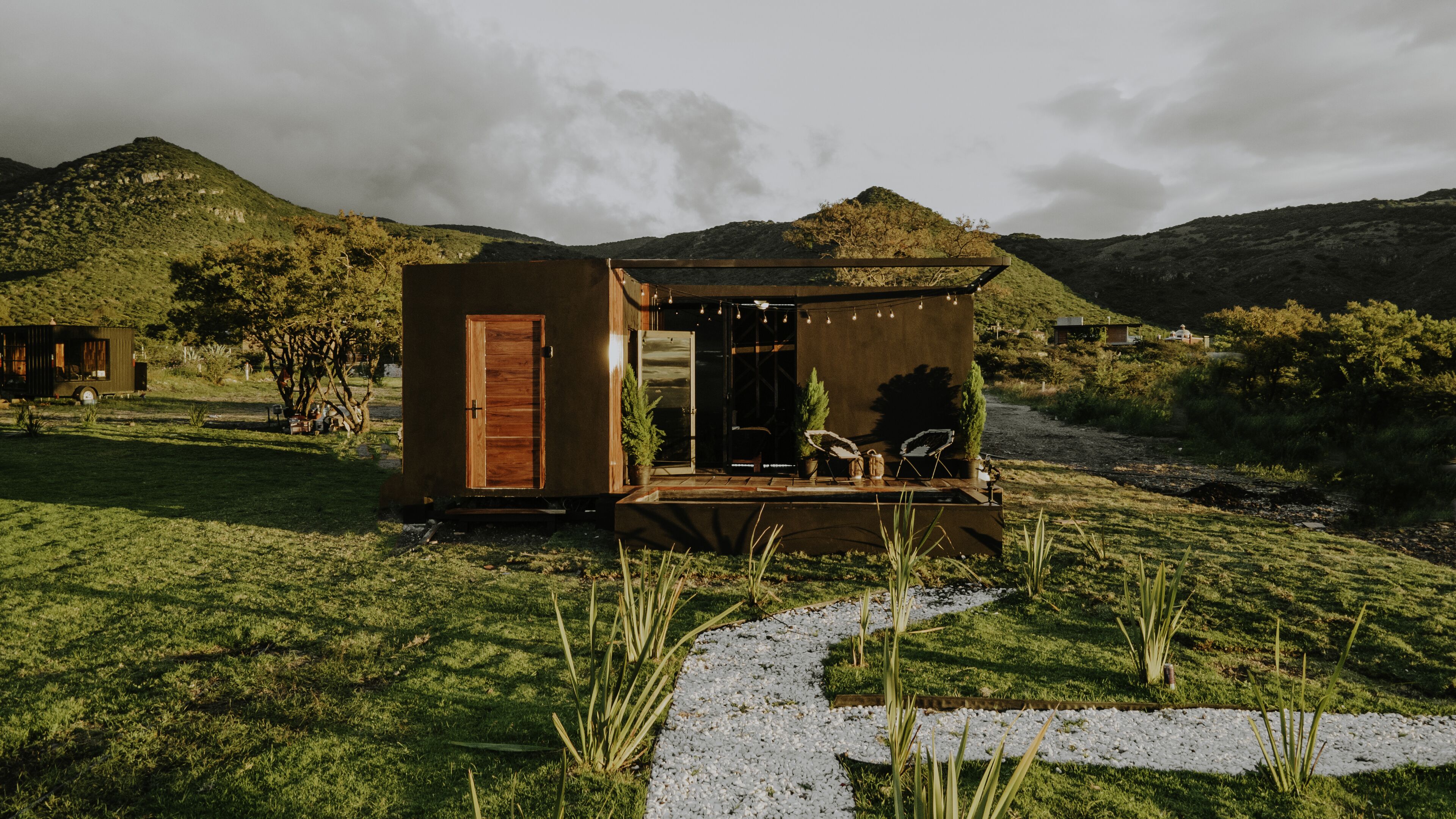 A small, wooden cabin with a door and a porch, surrounded by a grassy area and hills in the background.