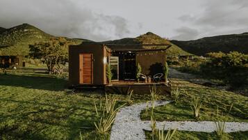A small, wooden cabin with a door and a porch, surrounded by a grassy area and hills in the background.