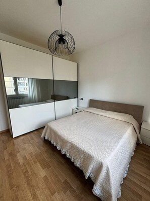 A bedroom with a large bed, a bedside table, a nightstand, a mirror, and a hanging light fixture.