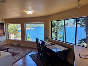 A room with a dining table and chairs, large windows with a view of a lake and forest, and a ceiling light fixture. - Shangrila Luxury Waterfront Panorama Suite | Oceanfront Seating & Beach Access (Pender Island)