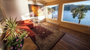 A modern living room with a large window overlooking a lake, a red sofa, a coffee table, and a potted plant. - Shangrila Luxury Waterfront Panorama Suite | Oceanfront Seating & Beach Access (Pender Island)