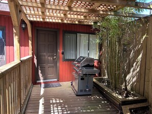 Una terraza de madera que conduce a un edificio rojo con una puerta y un ventanal. Una parrilla se encuentra ubicada en la terraza.