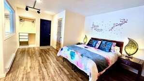 A bedroom with a wooden floor, a bed with a colorful bedspread, a nightstand with a lamp, and a wall with decorative elements. - Shangrila Luxury Waterfront Panorama Suite | Oceanfront Seating & Beach Access (Pender Island)