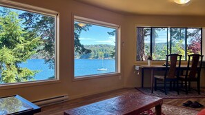 A room with large windows offering a view of a lake and forest, a wooden table, and a chair. - Shangrila Luxury Waterfront Panorama Suite | Oceanfront Seating & Beach Access (Pender Island)