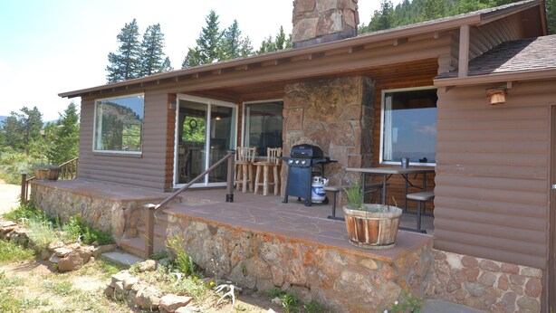 Une cabane en rondins avec une cheminée en pierre, un espace extérieur aménagé pour s’asseoir et une grande fenêtre donnant sur une zone boisée.