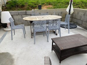 Um pátio externo com uma mesa redonda, quatro cadeiras, um banco de vime e um guarda-sol branco.