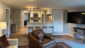 A modern living room with a brown leather sofa, a flat-screen TV, and a kitchen area with a bar stool. - An elegant and comfy Beaverton Home (Beaverton)
