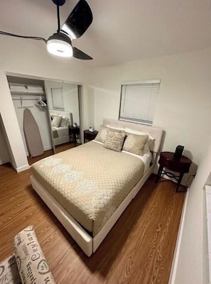 Quarto com cama, ventilador de teto, mesa de cabeceira, espelho e janela com persianas.
