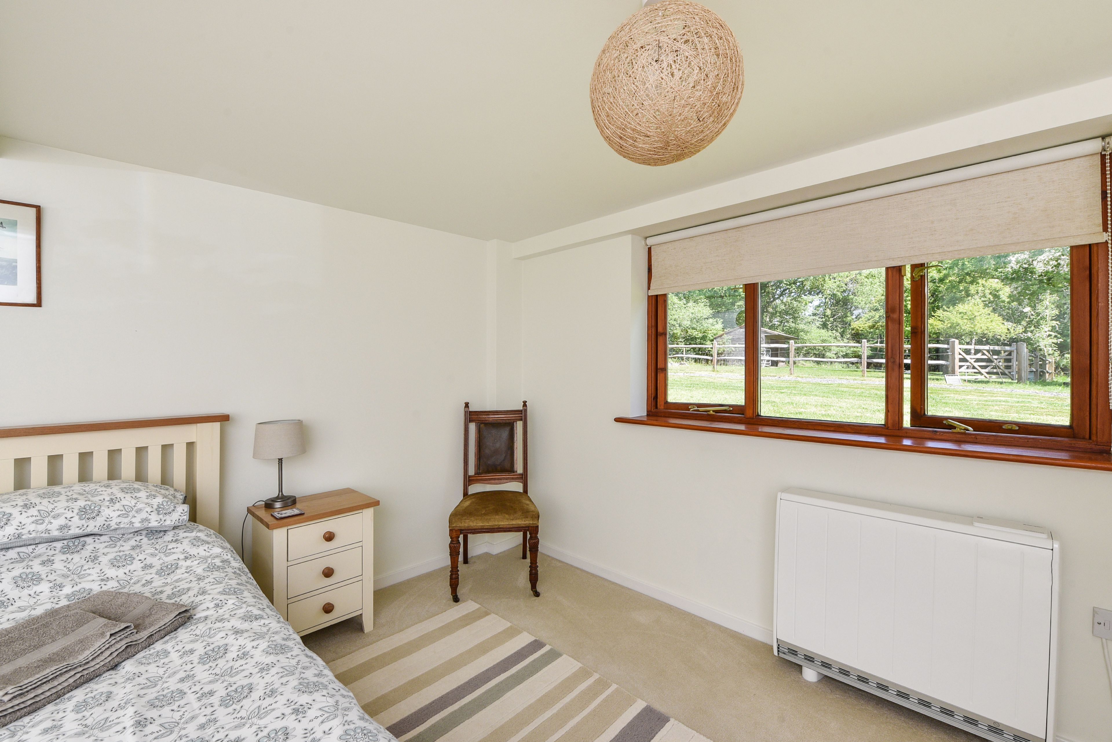 A bedroom with a bed, bedside table, chair, window, radiator, and a hanging light fixture.