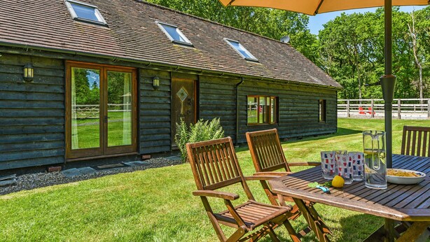 A log cabin with two windows, a door, and a patio with a table, chairs, and an umbrella.