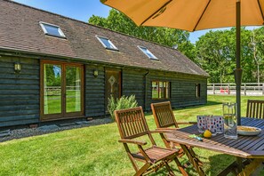 A log cabin with two windows, a door, and a patio with a table, chairs, and an umbrella. - Taylors Copse Cottage by inspirestays.com - a cottage that sleeps 5 guests in 2 bedrooms (Steep Marsh)