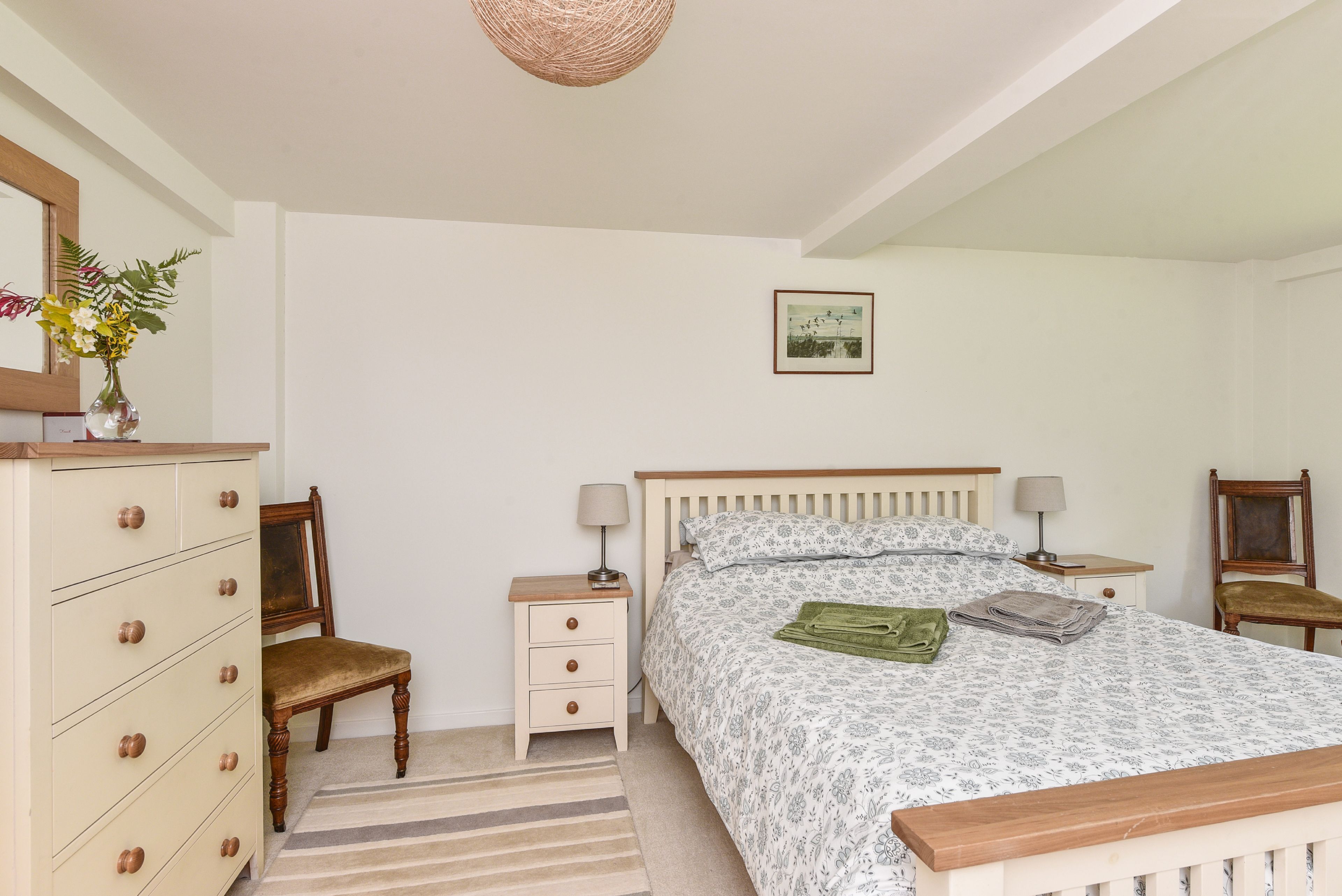 Une chambre à coucher comprenant un lit, une commode, une chaise, une table de chevet, une lampe et un tableau encadré accroché au mur.