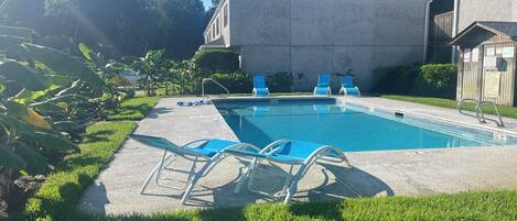 Una piscina con sillas azules, un deck de concreto y un edificio al fondo.