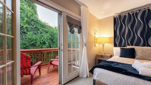Une chambre avec un lit, une table de chevet, une lampe et une vue sur une terrasse en bois et la verdure environnante.